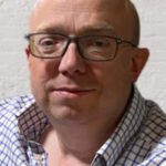 Headshot of Rob Ellis, a man wearing a white and blue checked shirt and dark framed glasses